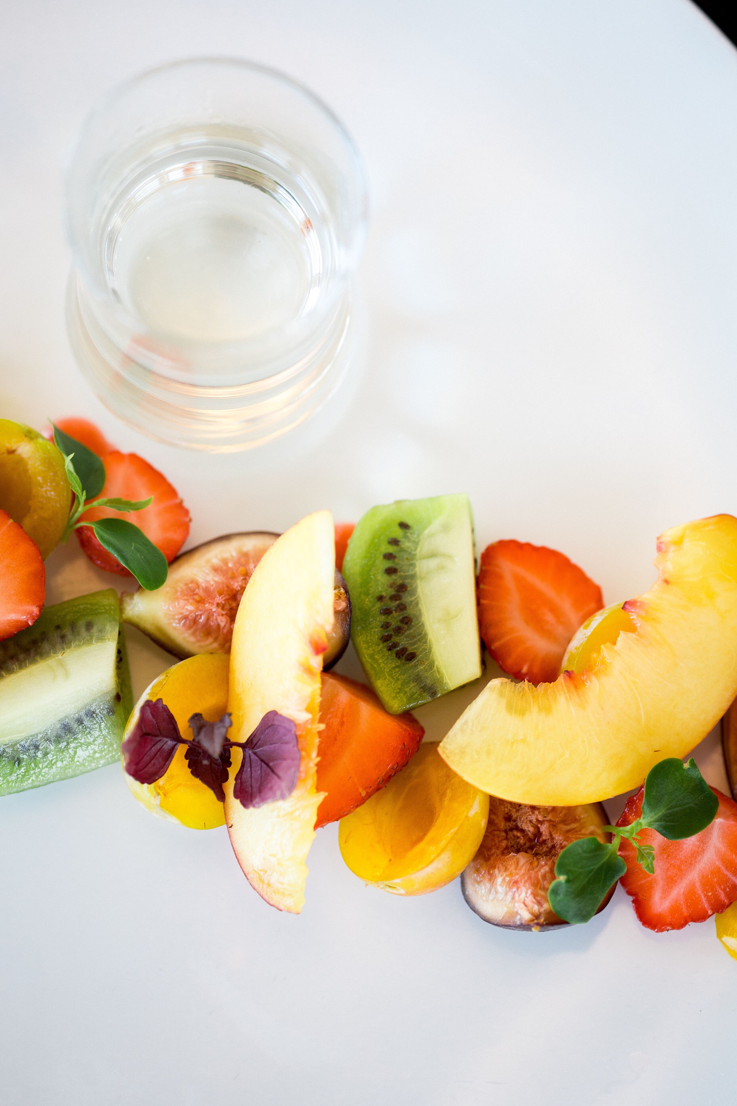 A variety of sliced fruits on a white plate, including kiwi, melon, and strawberries, with a small glass of water beside them.
