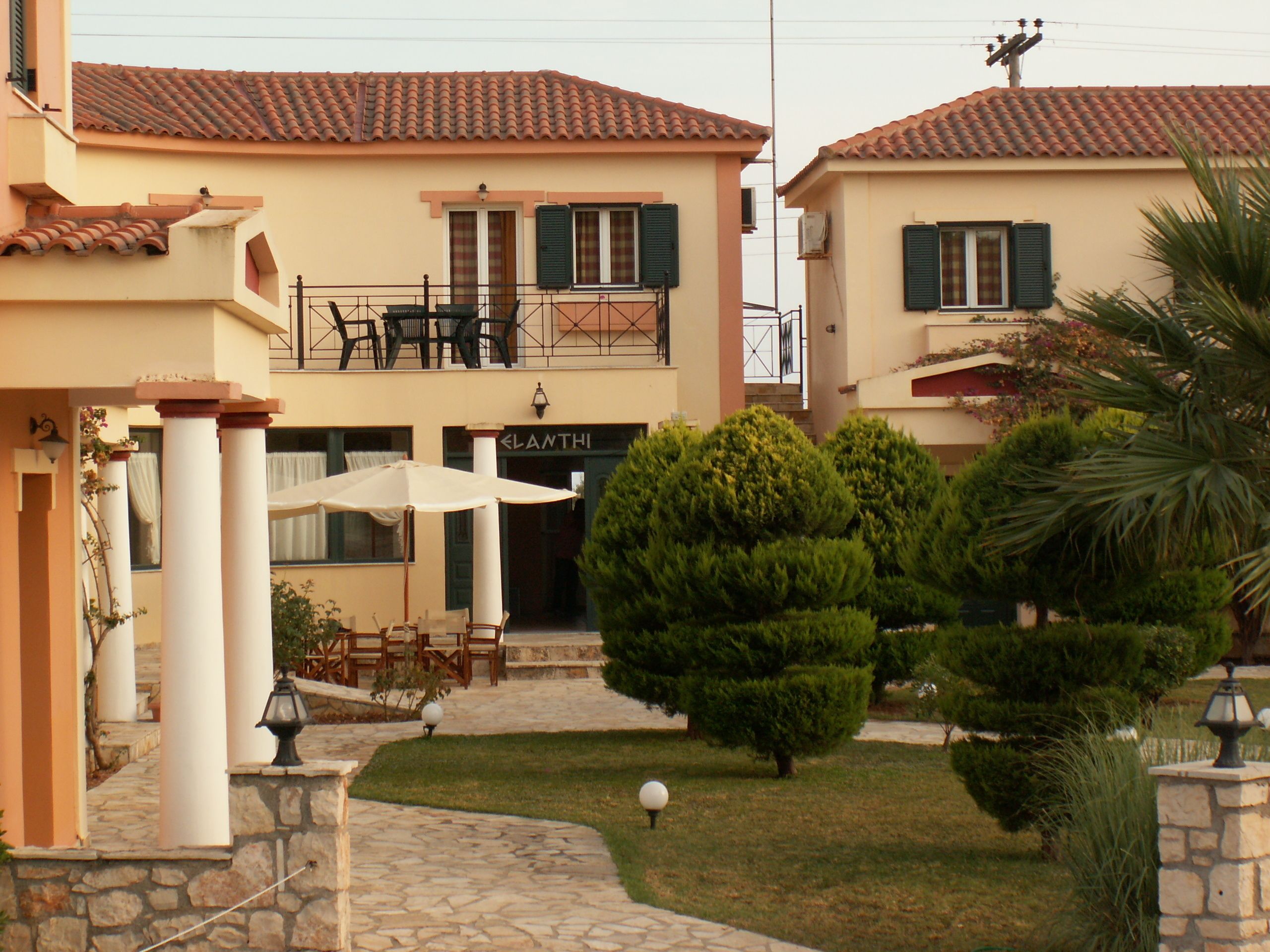 A building with a tiled roof, a balcony with outdoor furniture, and a well-maintained garden with trimmed bushes and a palm tree.