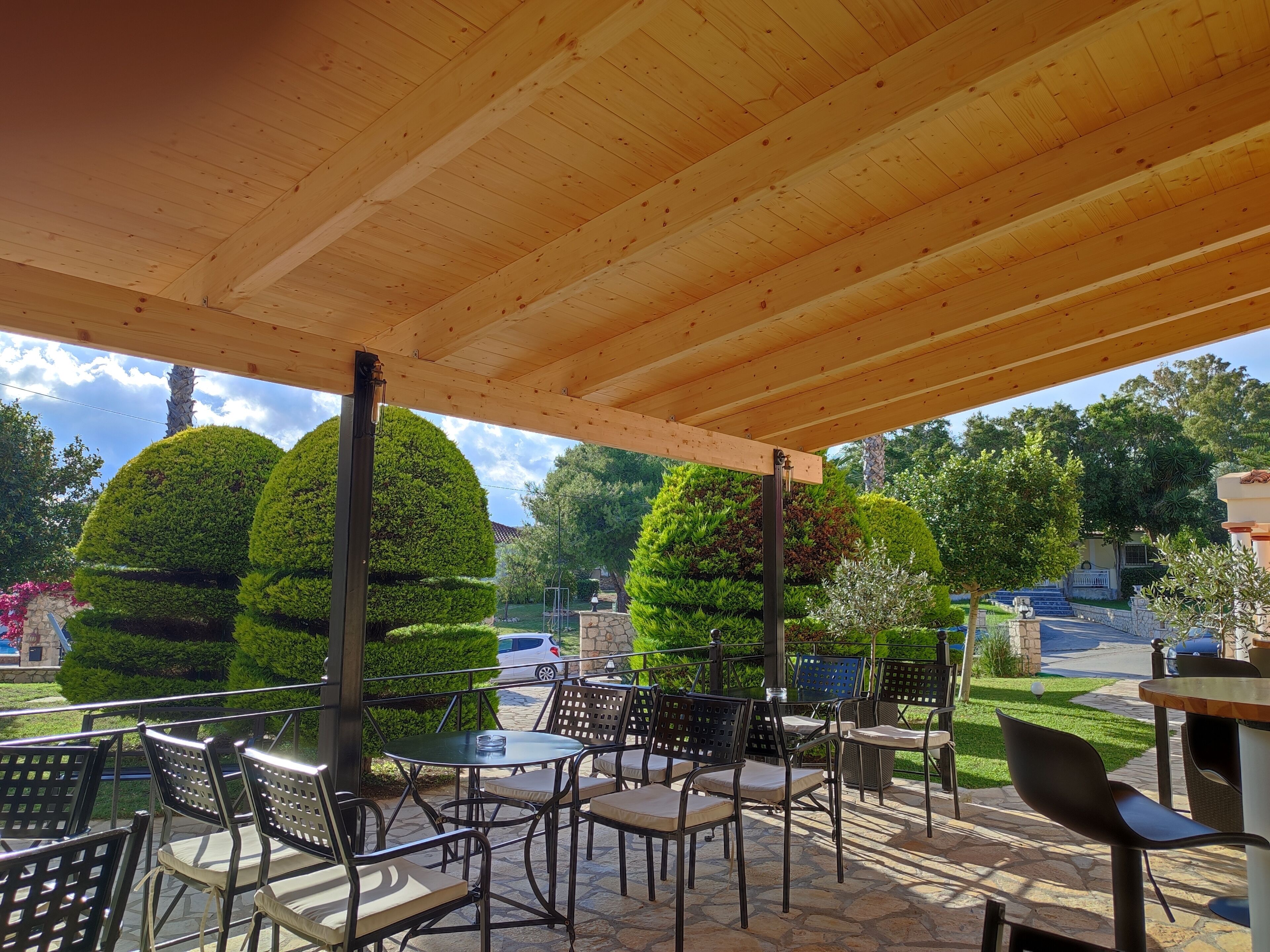 An outdoor patio area with round tables and chairs, surrounded by neatly trimmed trees and a stone pathway.