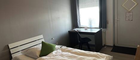 A bedroom with a bed, a desk, and a chair. There is a window with curtains, a door, and a wall-mounted light fixture.