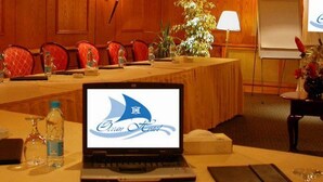 Une salle de conférence équipée d’un ordinateur portable affichant un logo, d’une longue table avec des chaises, des bouteilles d’eau et d’une petite table sur laquelle est posé un vase de fleurs.
