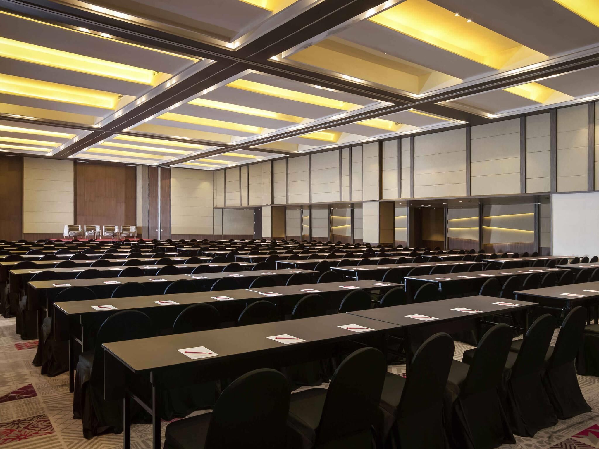 A conference room with rows of tables and chairs, a stage, and a modern ceiling design.
