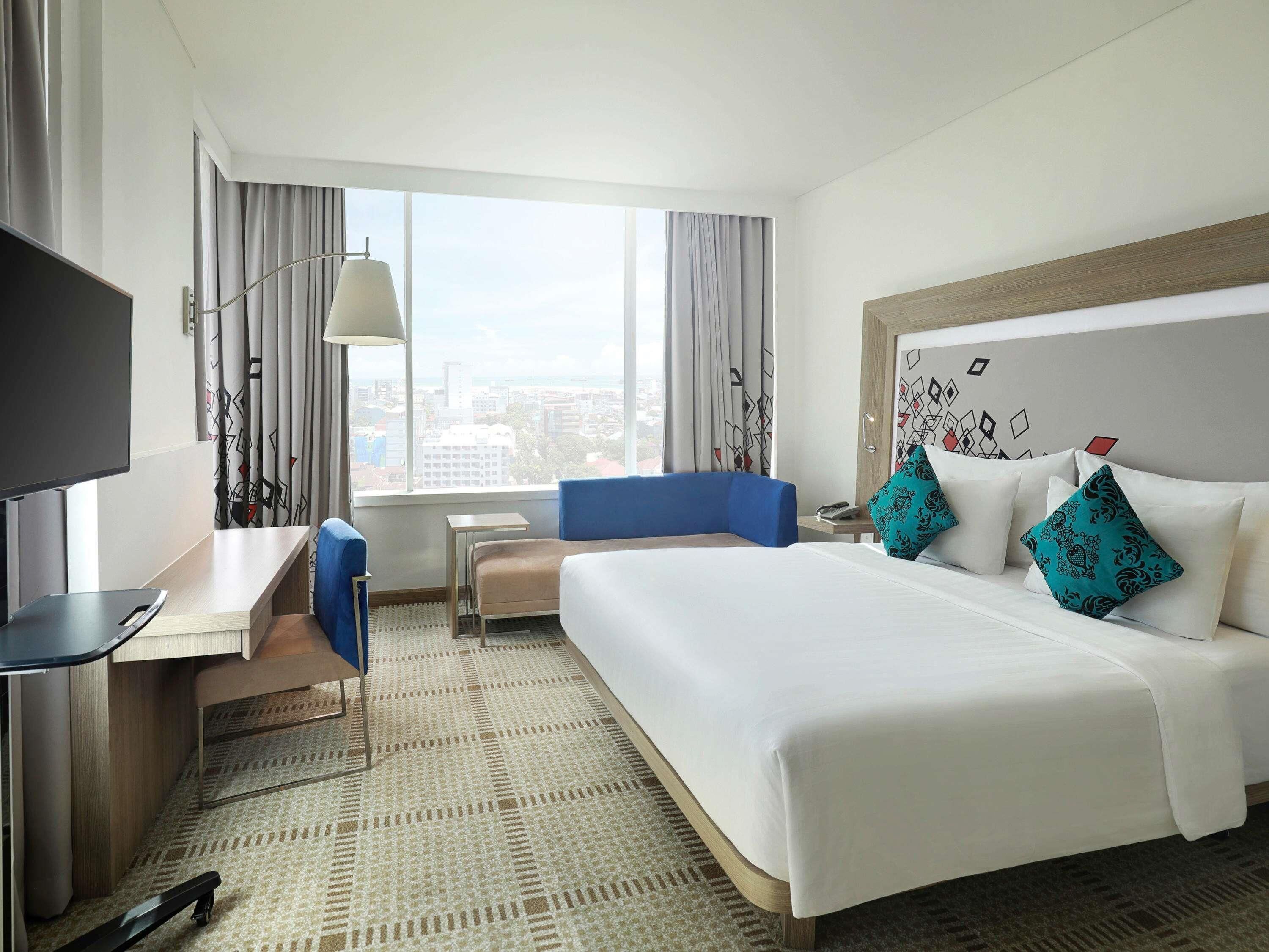 Habitación de hotel con cama, escritorio, silla, televisor y ventana con cortinas.