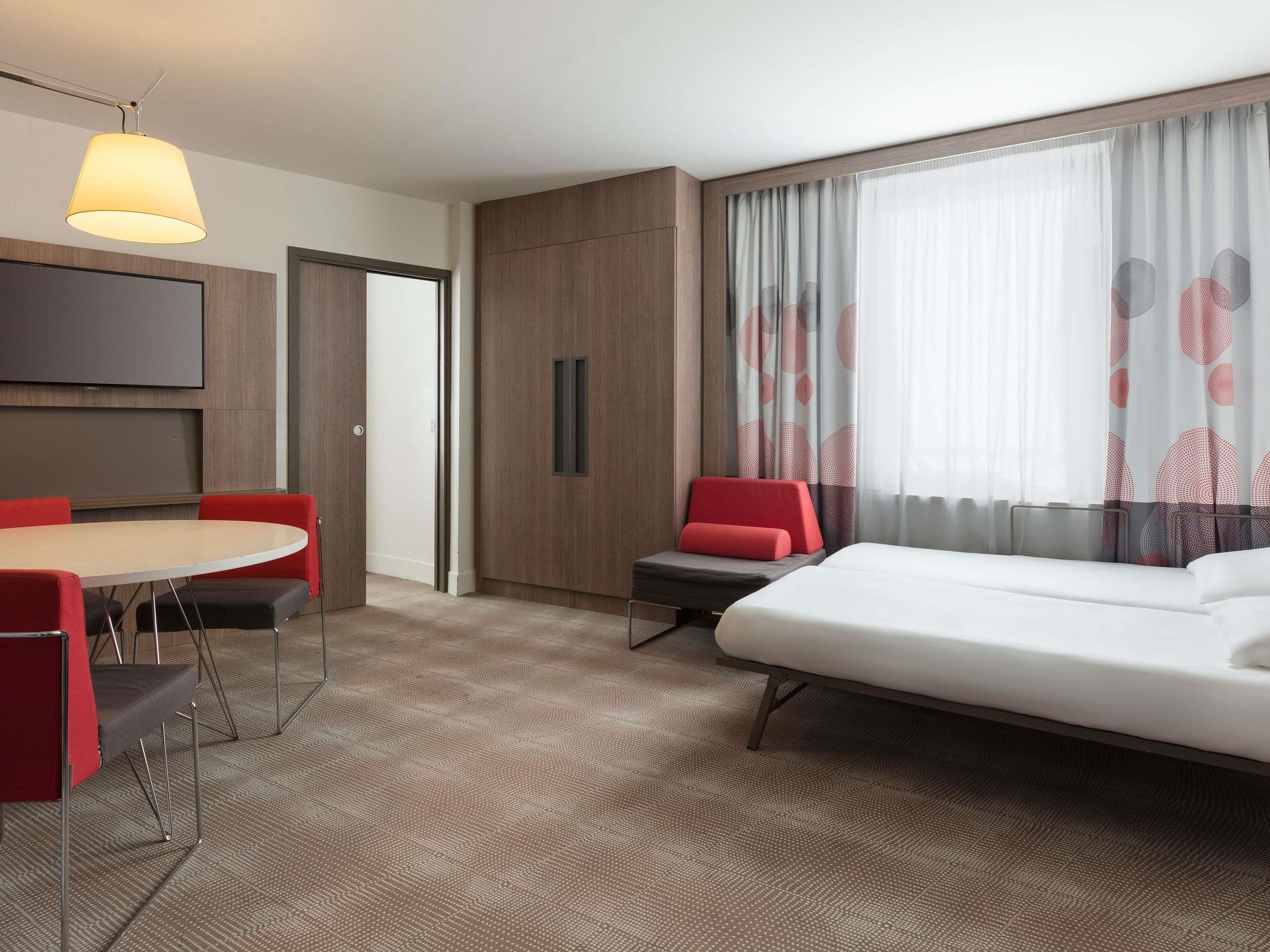 A hotel room with a bed, a dining table with chairs, a TV mounted on the wall, and a window with curtains.