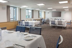 A conference room with tables set for a meeting, equipped with chairs, a projector screen, and a large window providing natural light.
