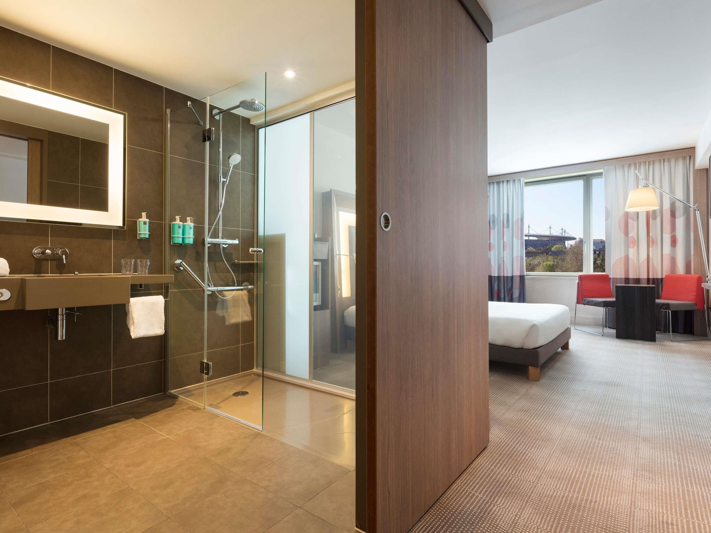 A modern hotel room with a bathroom featuring a glass shower enclosure, a vanity with a mirror, and a bed with a view of the city.