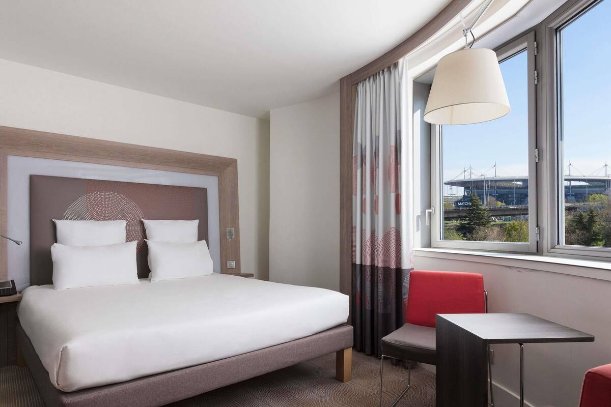 Habitación de hotel con cama, escritorio, silla, lámpara y ventana con vista a un puente.