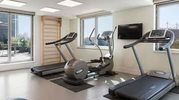 Un gimnasio con cintas de correr, elíptica y bicicleta estática, ventanas amplias y un televisor en la pared.