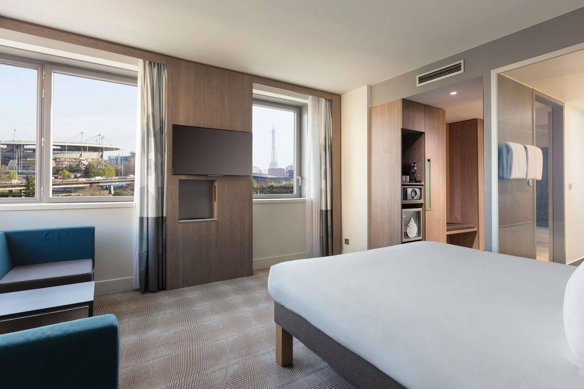 Habitación de hotel con cama, zona de estar, televisor, armario y vistas a la ciudad.