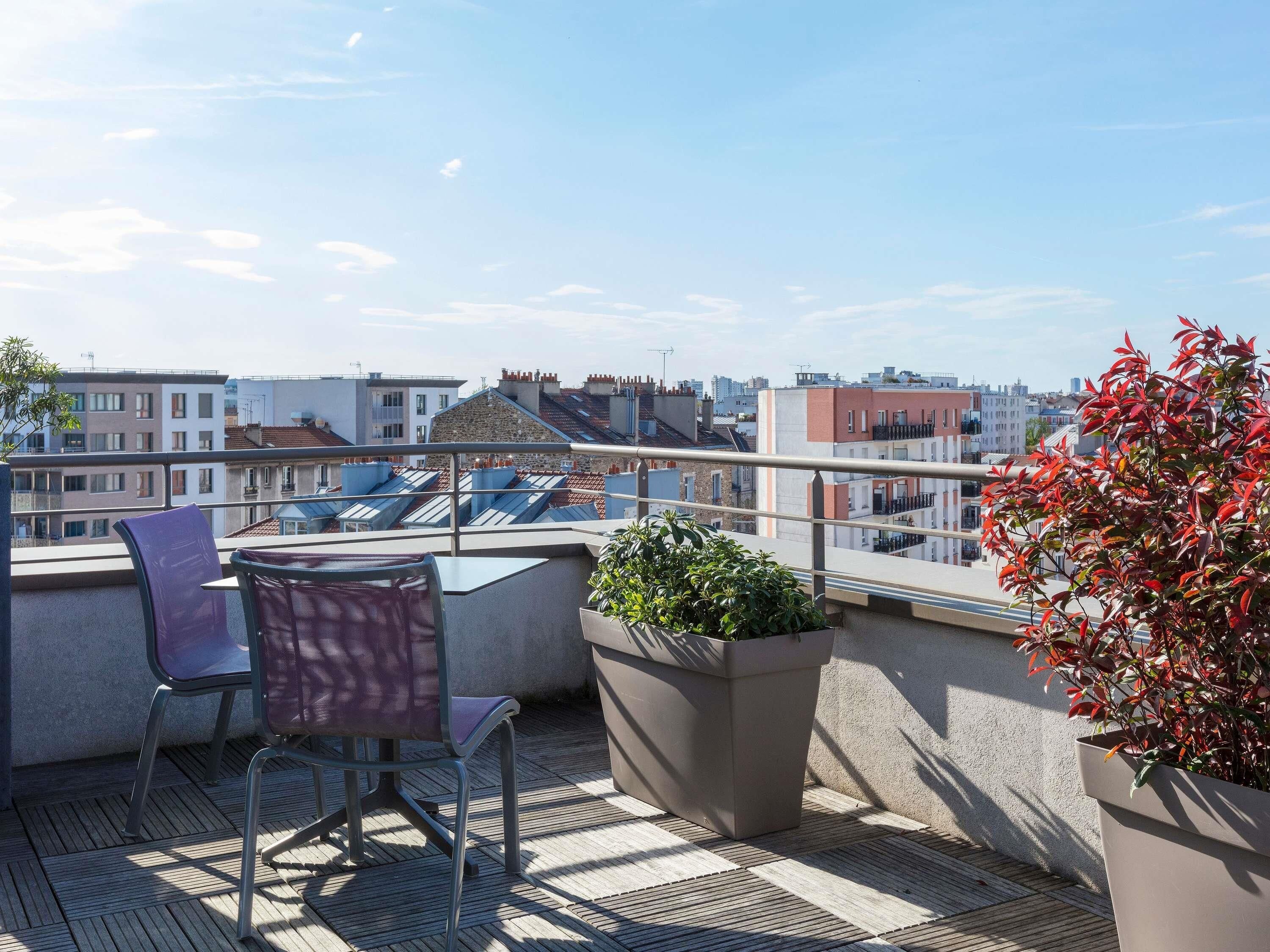A rooftop terrace with a table and chairs, a potted plant, and a view of the cityscape.