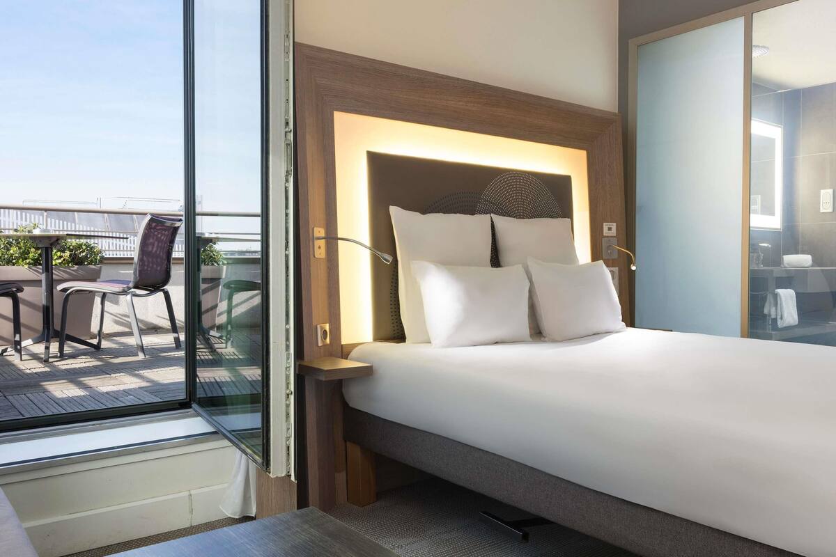 Una habitación de hotel moderna con una cama grande, una mesita pequeña y una silla. Tiene un balcón con vistas a la ciudad.