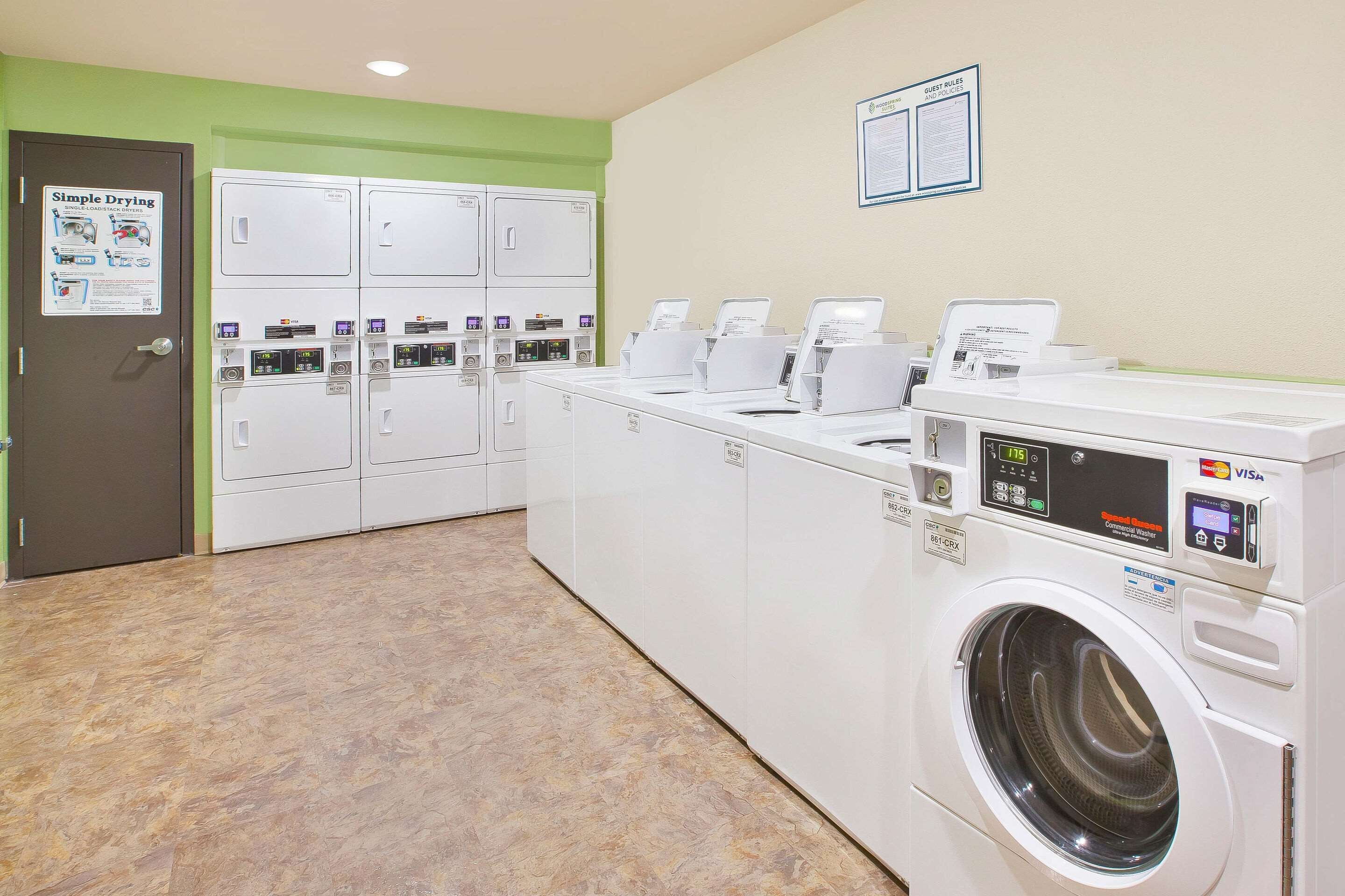 Une buanderie équipée de plusieurs machines à laver et de plusieurs sèche-linge, une affiche simple indiquant les instructions de séchage et un panneau affichant les règles à respecter par les clients.