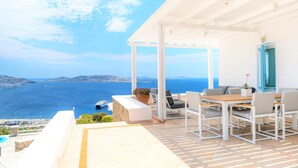 A terrace with a view of the sea, a wooden table, and white chairs. - Aeri Villas & Apartments (Mykonos)
