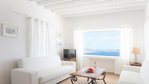 A white living room with a sofa, a coffee table with a flower vase, a TV, and a window with a view of the sea. - Aeri Villas & Apartments (Mykonos)