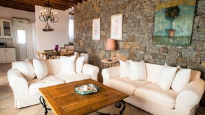 A living room with two white sofas, a wooden coffee table, a stone wall, and a kitchen in the background. - Aeri Villas & Apartments (Mykonos)