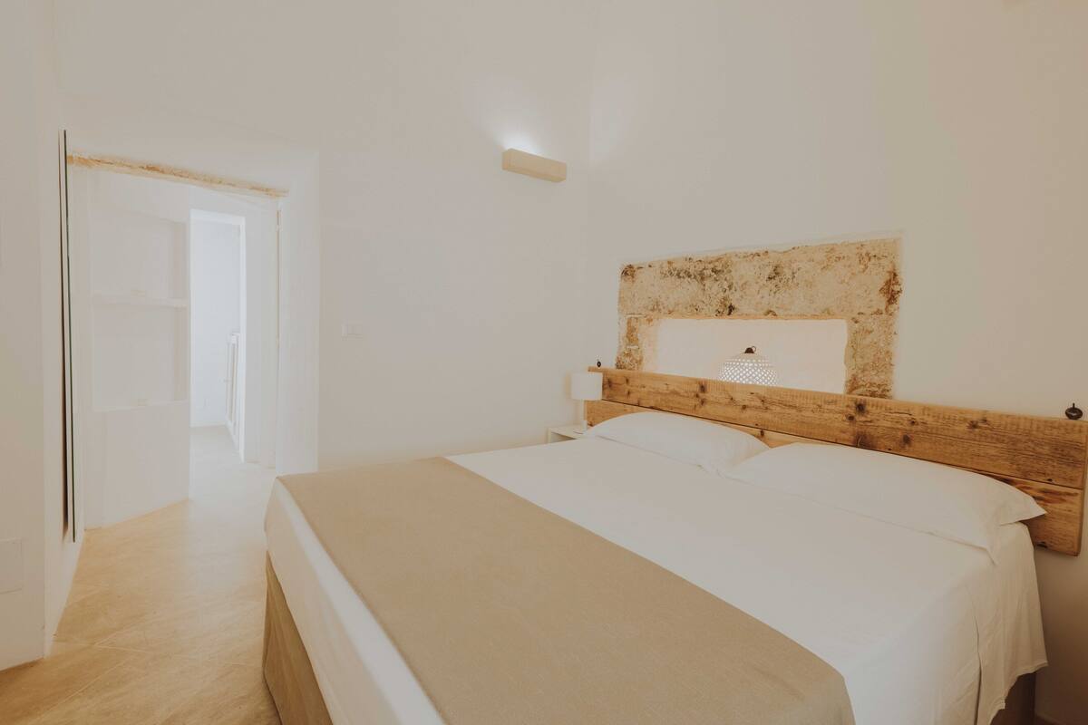 Quarto com cabeceira de madeira, cama com roupa de cama branca e porta que dá para outro cômodo.