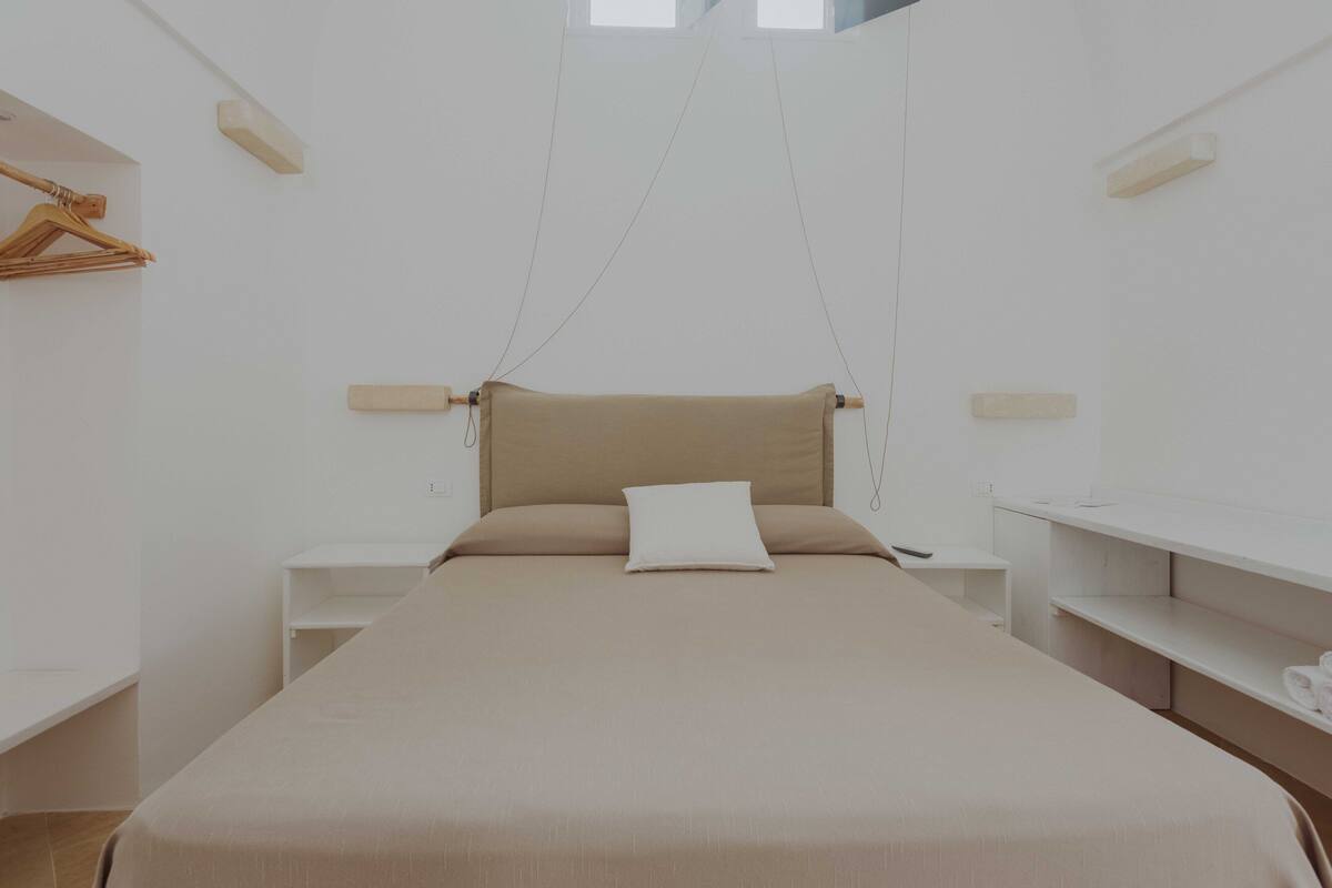 Quarto minimalista com uma cama ao centro, ladeada por mesas de cabeceira brancas e um pendente.