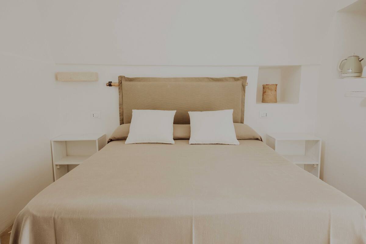 Uma cama com cabeceira bege e duas almofadas brancas, com mesas de cabeceira brancas de cada lado, cada uma com uma pequena prateleira.