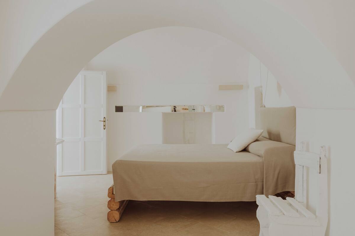 Quarto minimalista com cama grande, porta em arco e guarda-roupa branco.