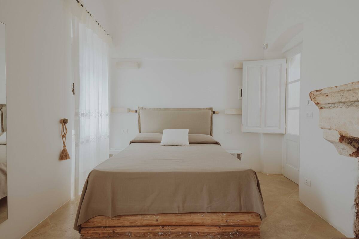 Quarto com cama, um banco de madeira e parede branca com detalhe em tijolos.