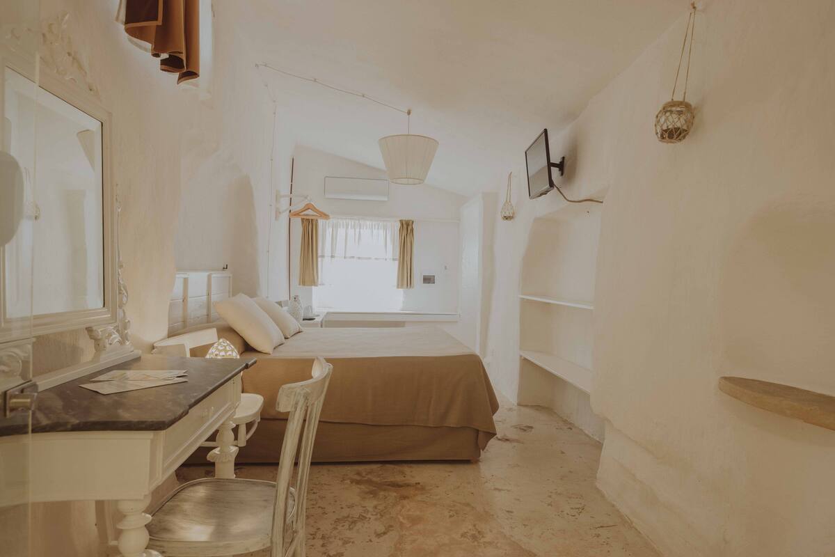 Quarto com cama, escrivaninha e cadeira. Há uma janela com cortinas, luminária pendente e prateleira na parede.