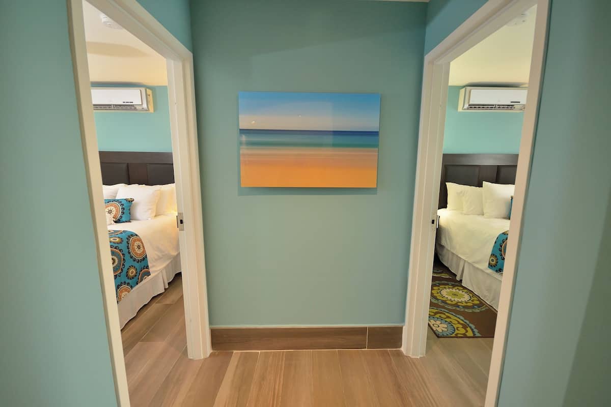 A hotel room with a large painting of a beach scene on the wall, a bed with a patterned bedspread, and an air conditioning unit above the bed.