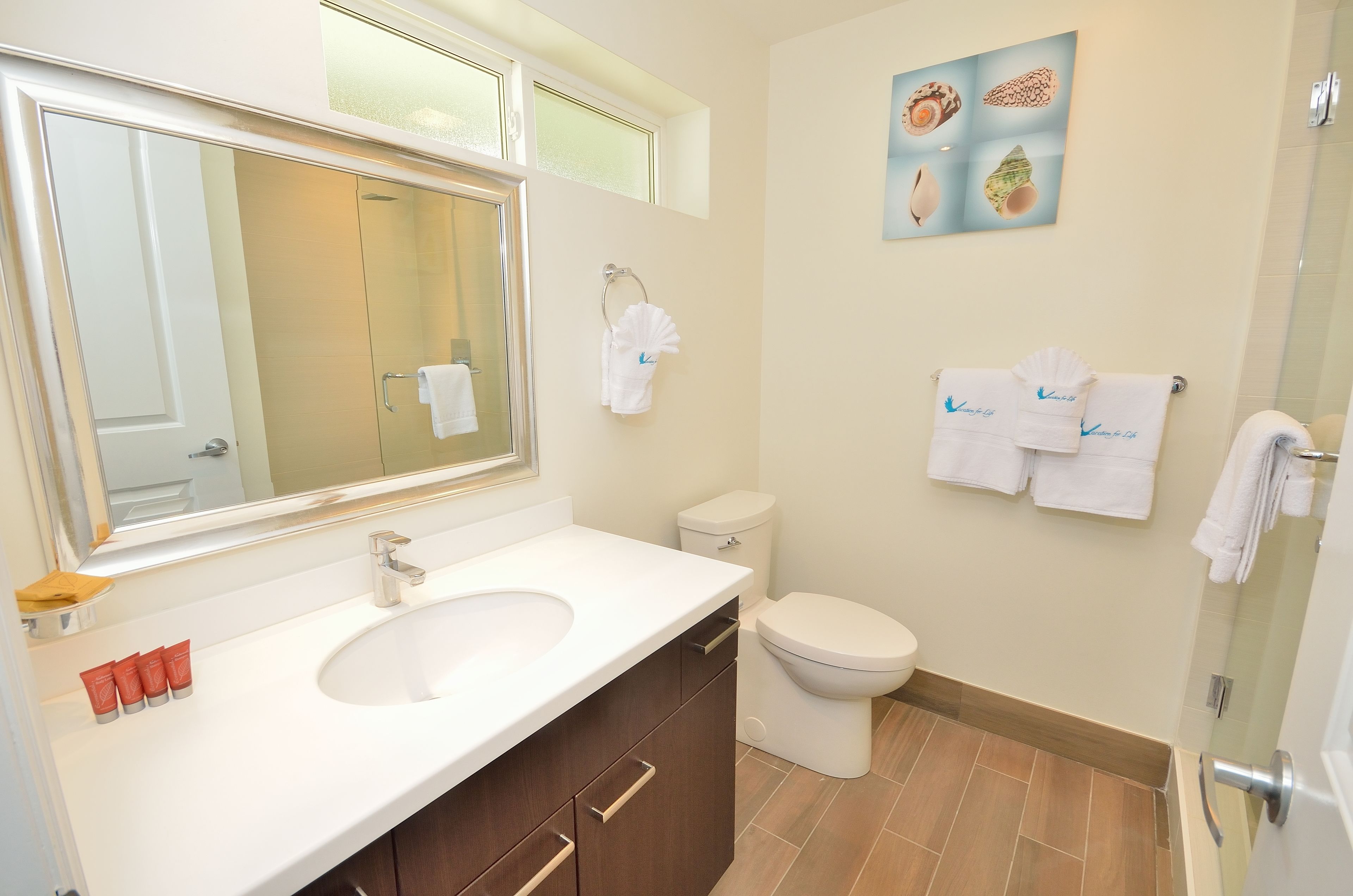 A bathroom with a large mirror, a sink, a toilet, and a shower. There are towels hanging on the wall and a framed picture above the toilet.