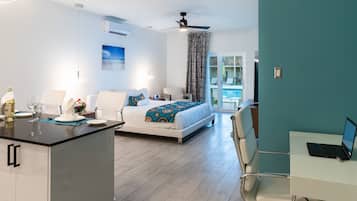 A modern hotel room with a bed, a desk with a laptop, a dining area with a table and chairs, and a view of the pool.