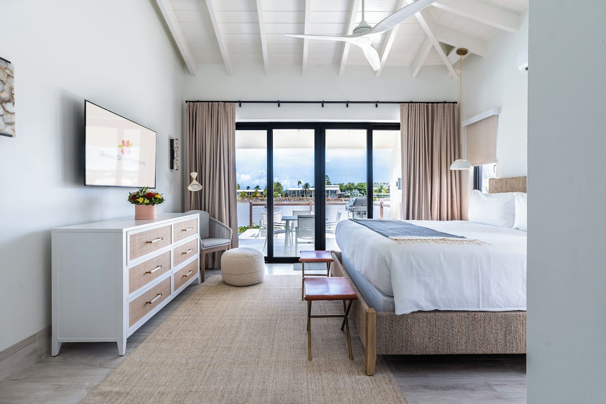 A modern hotel bedroom with a large bed, a dresser, a bench, and a view of the outdoors.