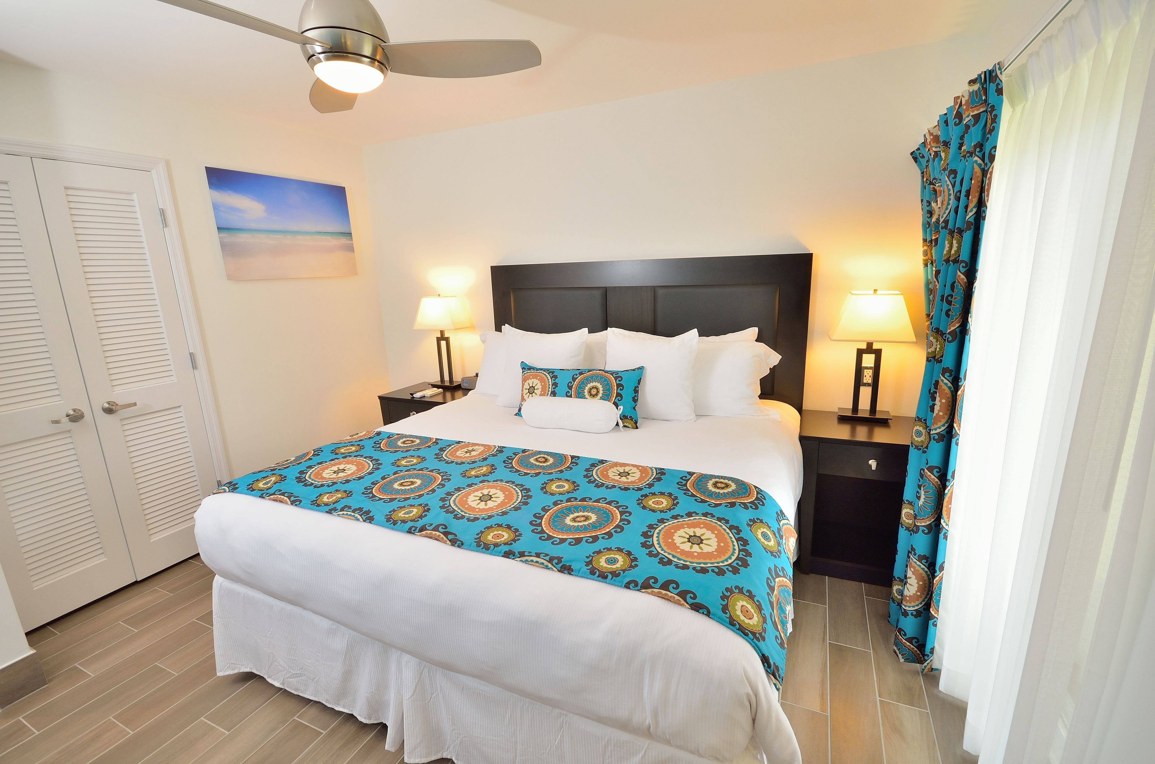Une chambre d’hôtel bien aménagée, avec un lit, deux tables de chevet, un ventilateur de plafond et une vue sur l’océan.
