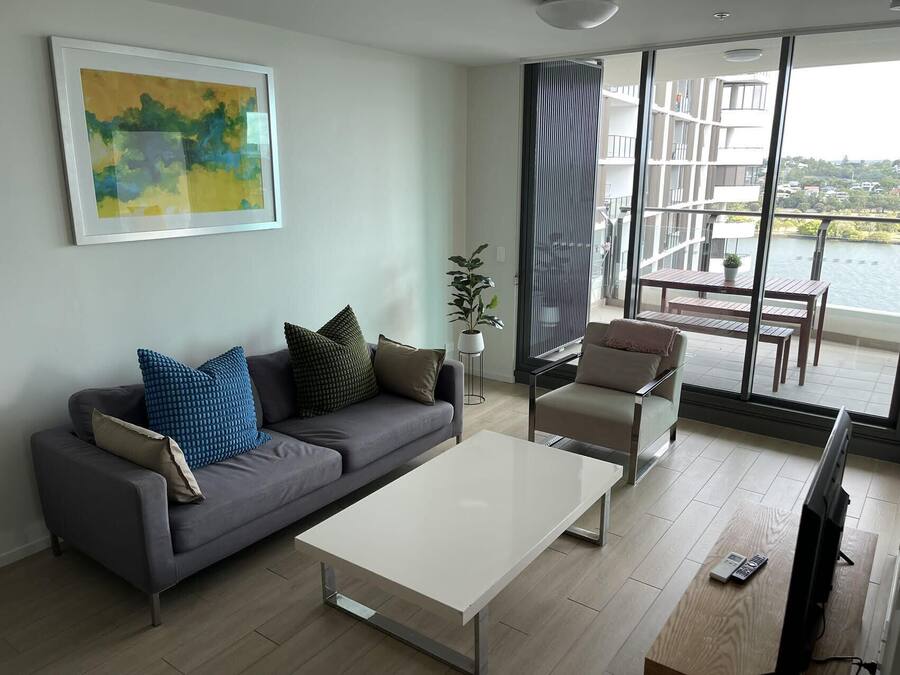 A modern living room with a grey sofa, a white coffee table, and a beige armchair. There is a large window with a view of a balcony and buildings.