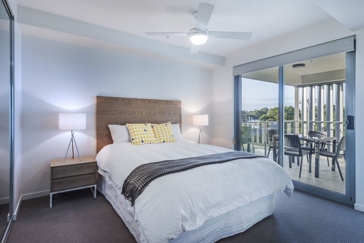 A modern hotel room with a large bed, two bedside lamps, a ceiling fan, and a balcony with outdoor furniture.