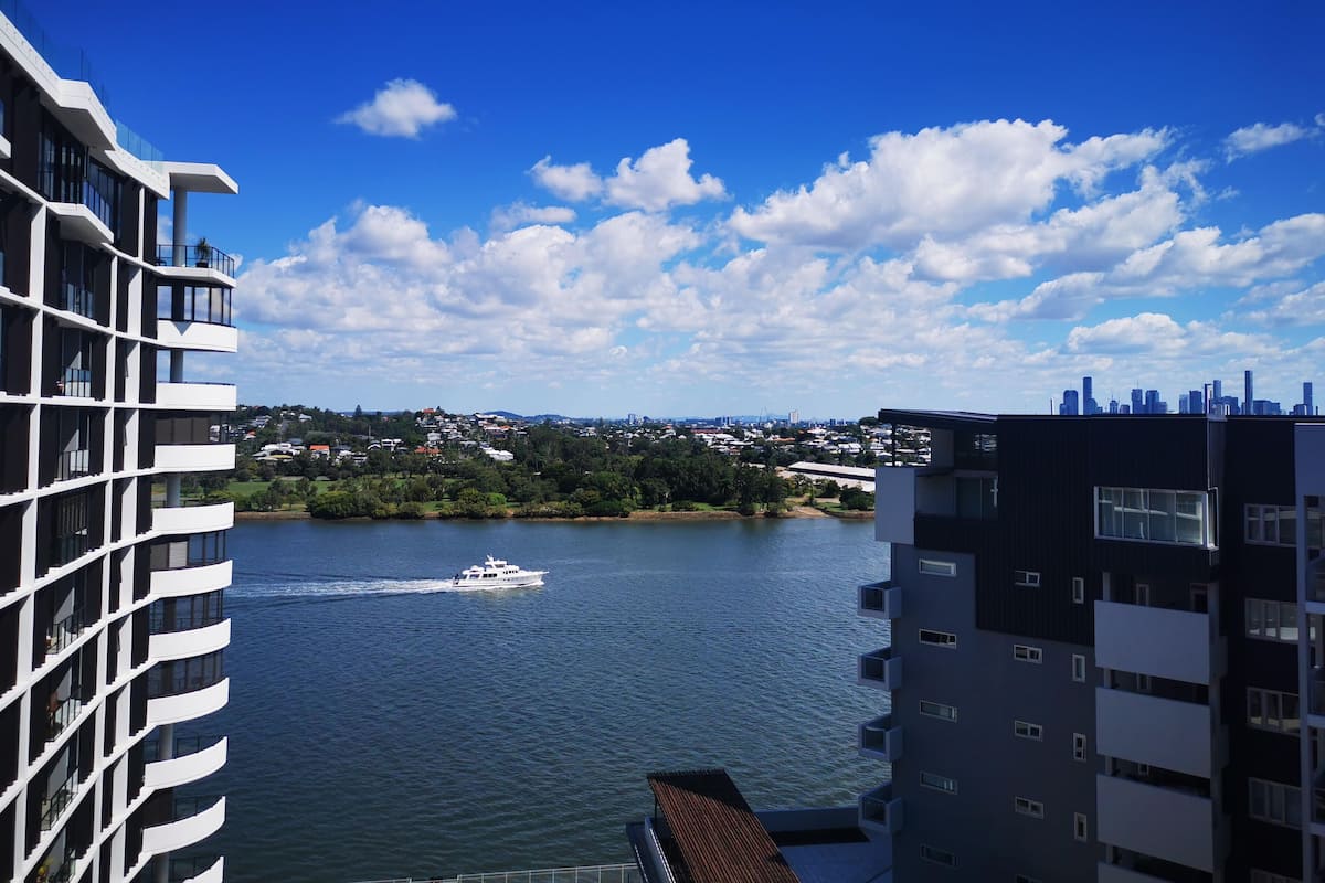 A modern building with balconies overlooking a body of water, a yacht on the water, and a cityscape in the distance.