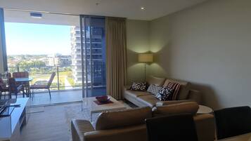 Una sala de estar moderna con sofá, mesa de centro y lámpara. Hay un balcón con vistas a edificios y una mesa con sillas.