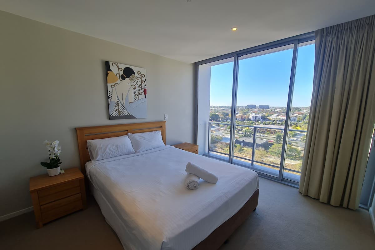 A hotel room with a large bed, a nightstand with a plant, a painting on the wall, and a view of the city through large windows.