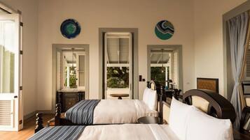 A bedroom with two beds, a small table, and a chair. There are two windows with shutters, and a view of greenery outside.