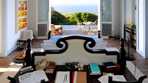 Un dormitorio con una cama grande, un escritorio con libros y una lámpara, y vista al océano a través de una puerta abierta.