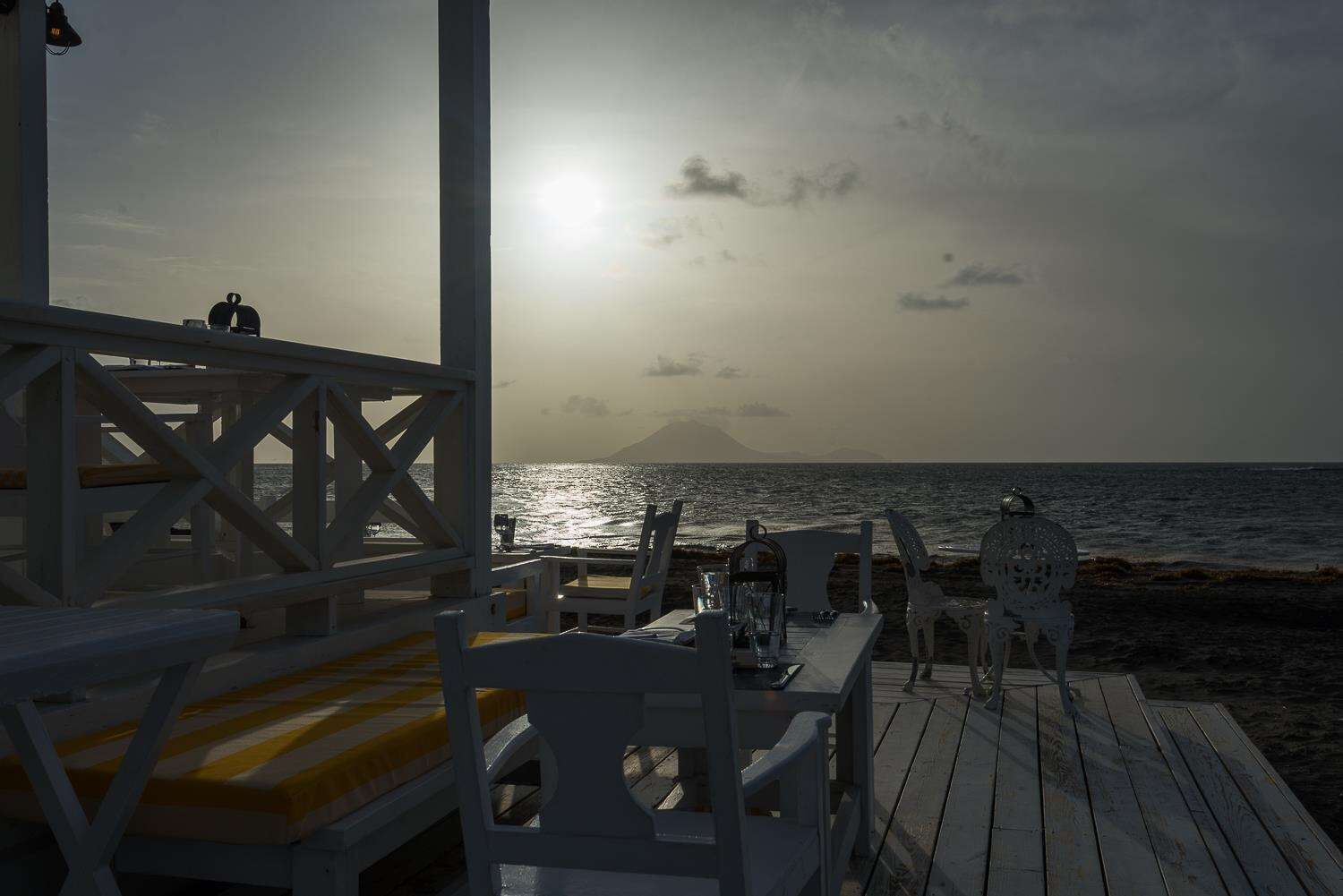 A wooden deck with white chairs and tables, overlooking the sea with a sunset in the background.
