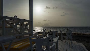 A wooden deck with white chairs and tables, overlooking the sea with a sunset in the background.