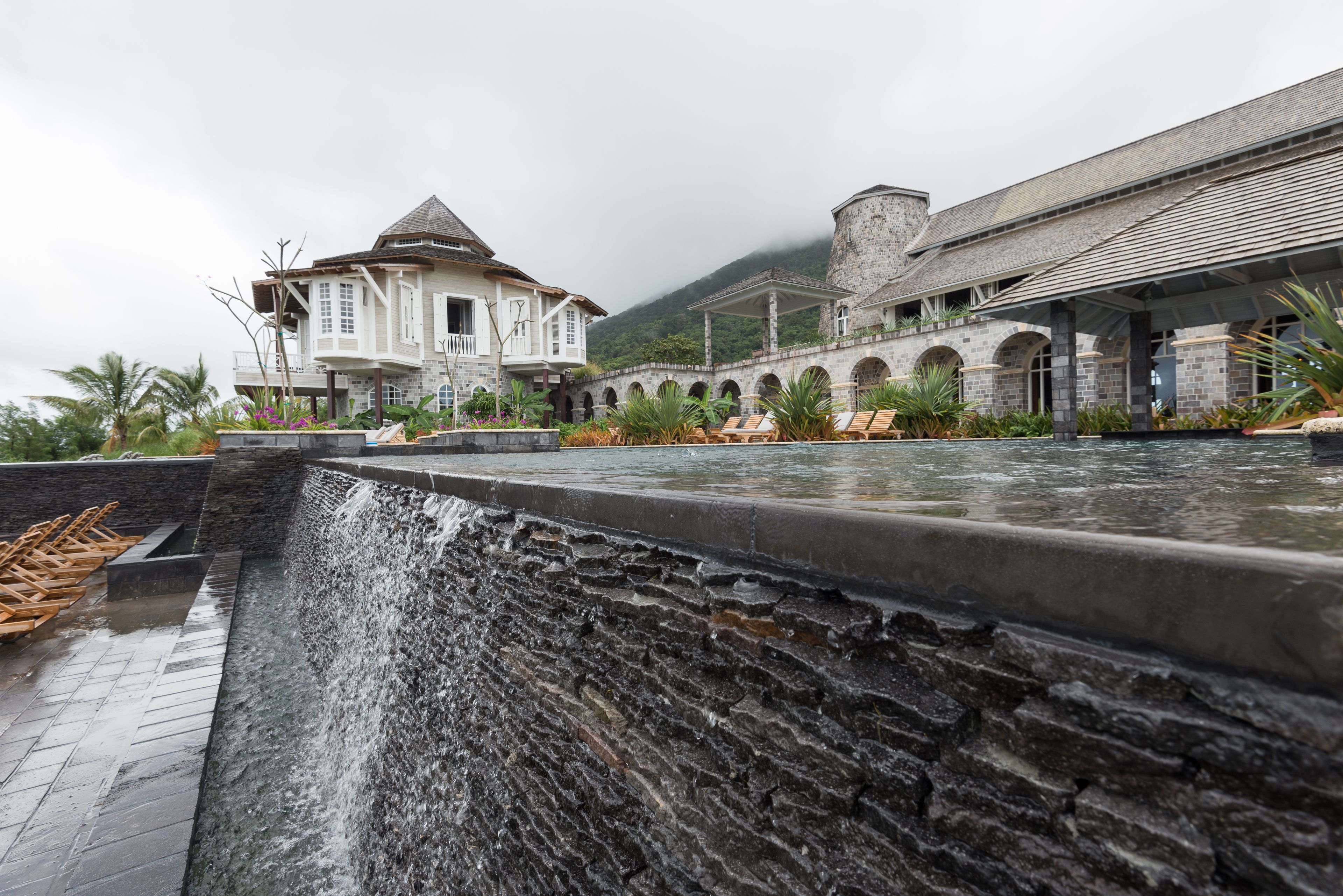 A stone water feature with a waterfall, a pool with lounge chairs, and a building with a stone facade and arched windows.