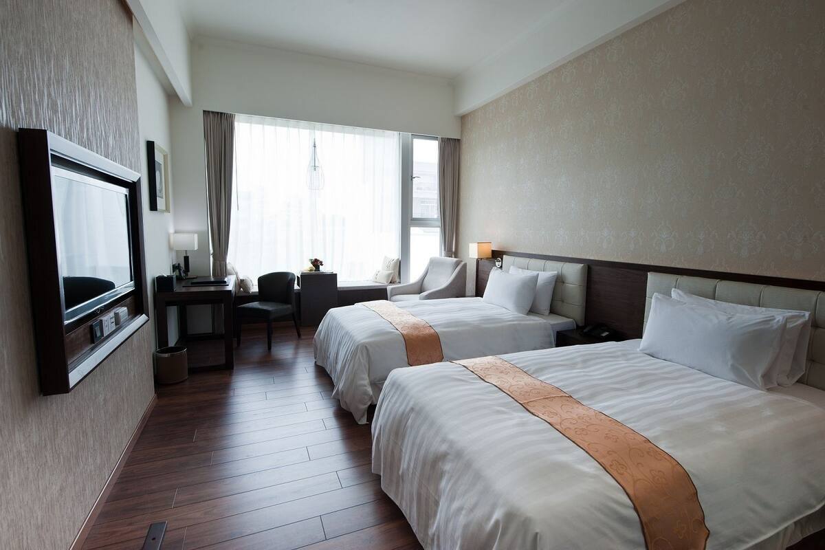 A hotel room with two beds, a desk, a chair, a TV, and a window with curtains.