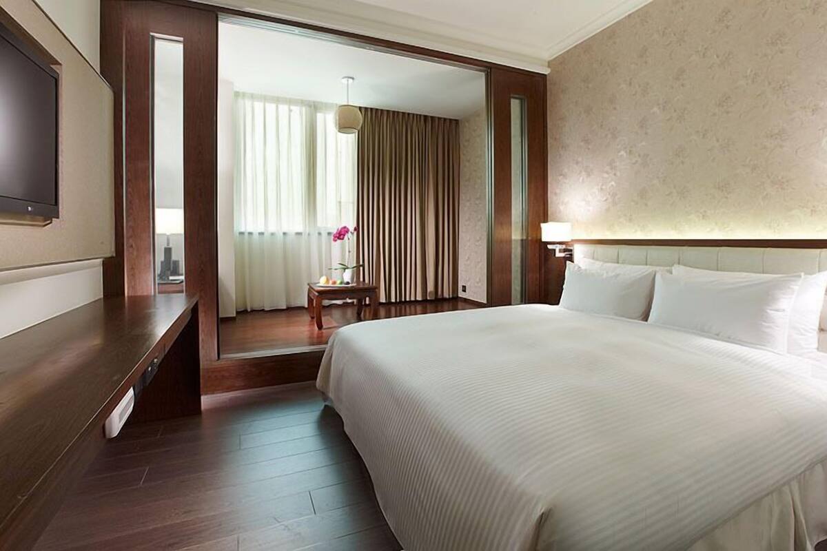 A hotel room with a large bed, a TV mounted on the wall, a desk with a chair, and a view of the city through the window.