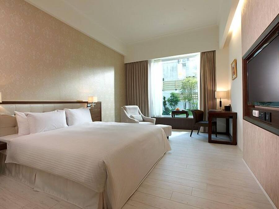 Una habitación de hotel con una cama grande, televisión, una silla y un ventanal con cortinas.