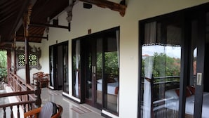 A covered outdoor seating area with wooden chairs and a table, large windows with curtains, and a view of greenery and a building in the background. - Taman Agung Hotel (Denpasar)