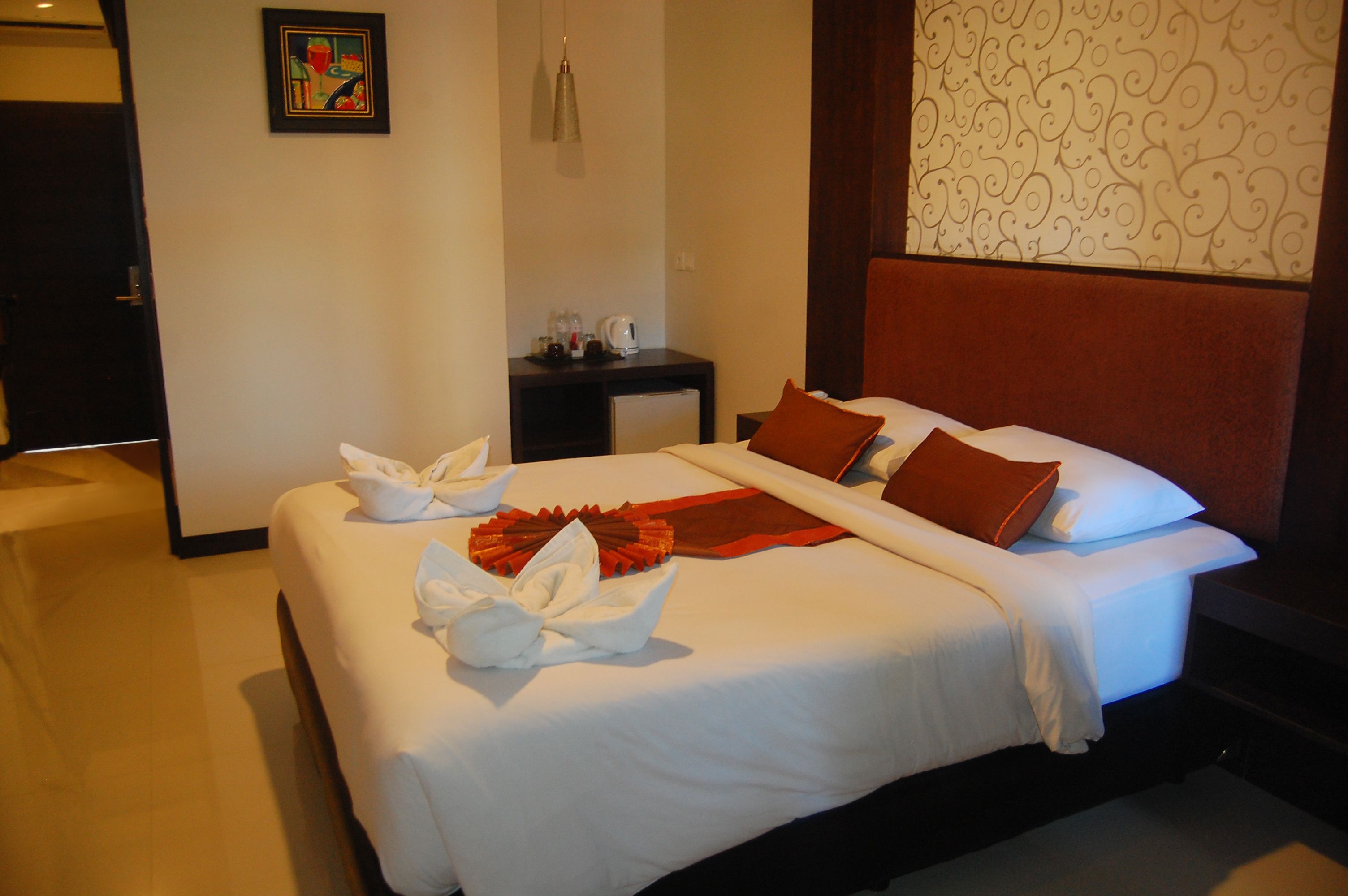 A neatly made bed with white linens and a red throw blanket, a small bedside table with a lamp, and a wall with a decorative pattern.