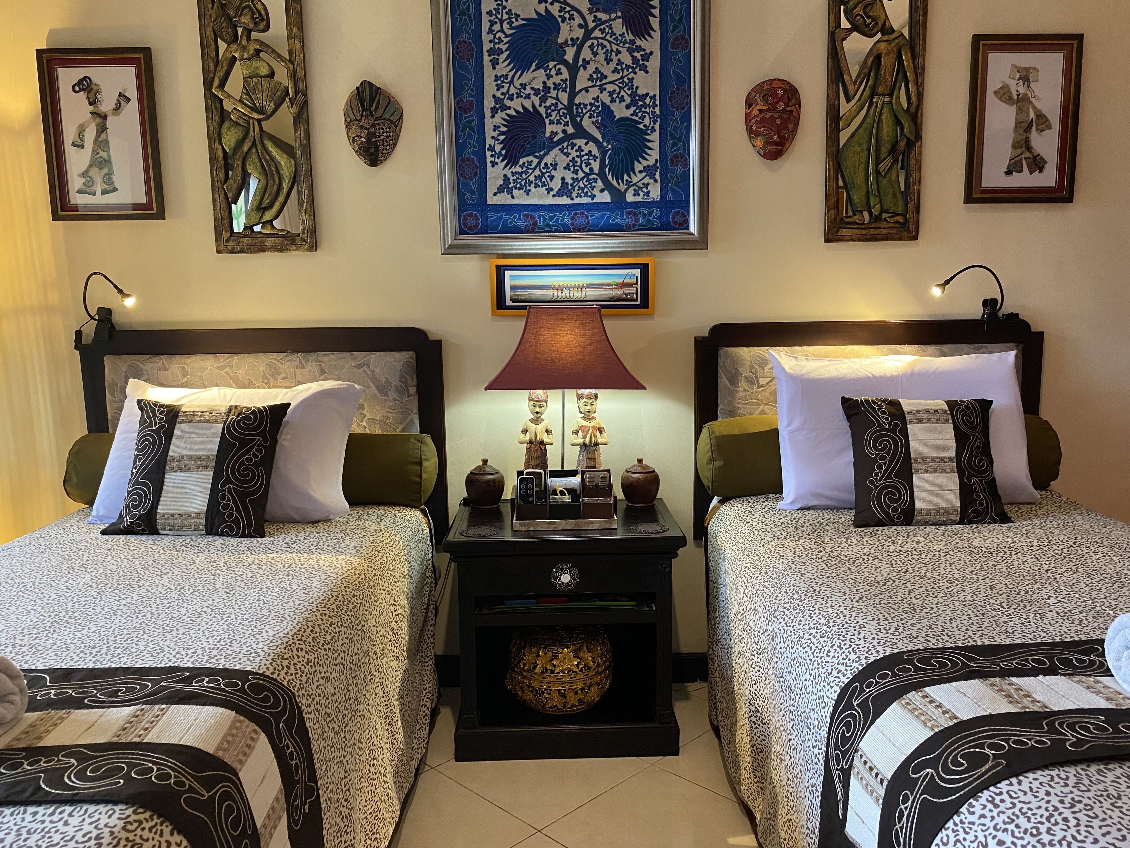 A hotel room with two beds, each with patterned bedding and decorative pillows. There is a nightstand with a lamp and a small statue between the beds. The walls are adorned with framed artwork and masks.