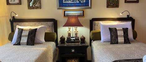 A hotel room with two beds, each with patterned bedding and decorative pillows. There is a nightstand with a lamp and a small statue between the beds. The walls are adorned with framed artwork and masks.