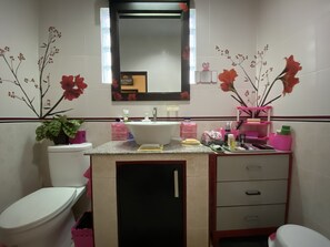 A bathroom with a white sink, a mirror above it, and a toilet. There are pink storage boxes and a potted plant on the counter. - Taman Agung Hotel (Denpasar)