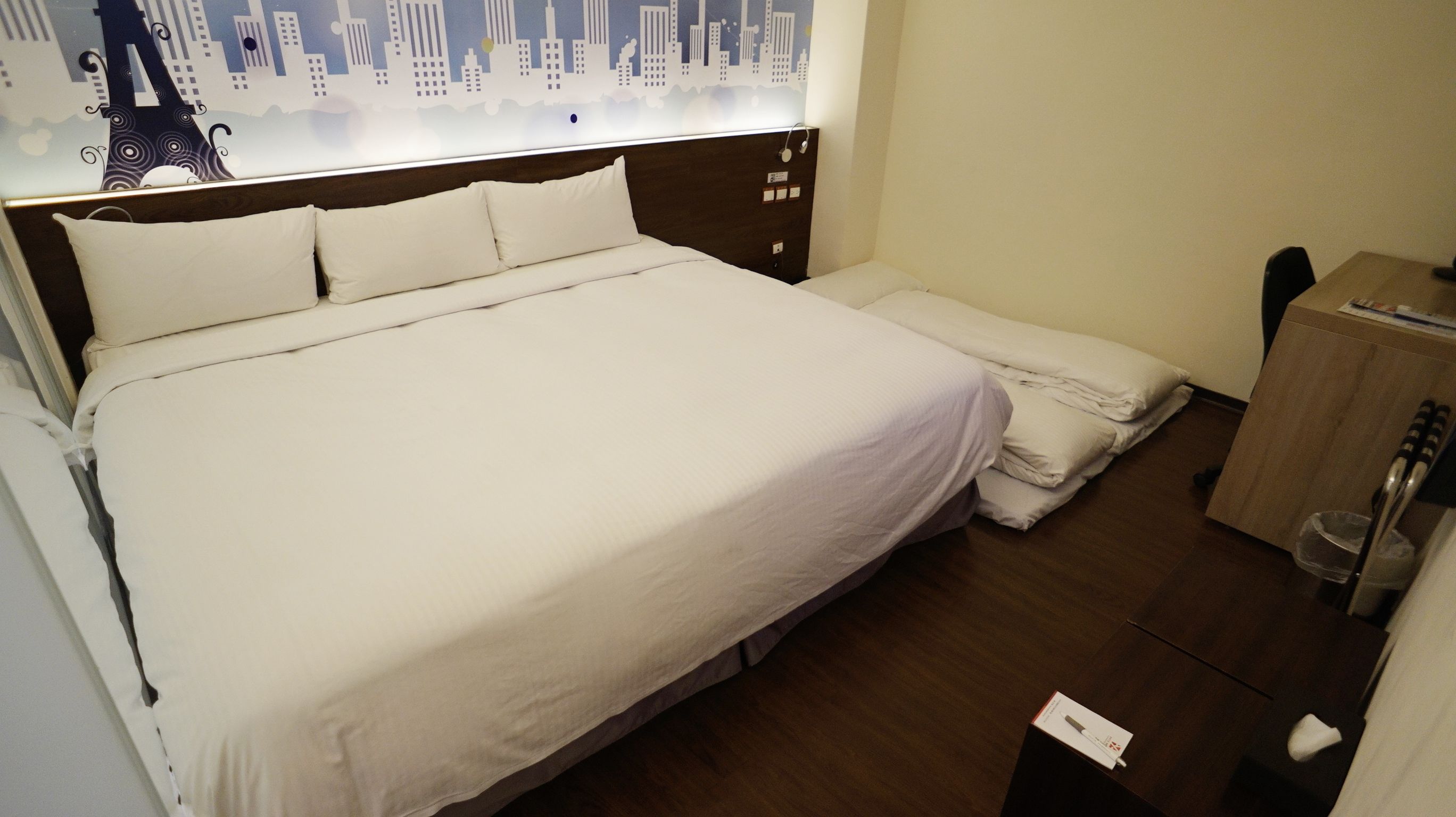 A hotel room with a large bed, a wooden headboard, a nightstand, and a view of a cityscape.