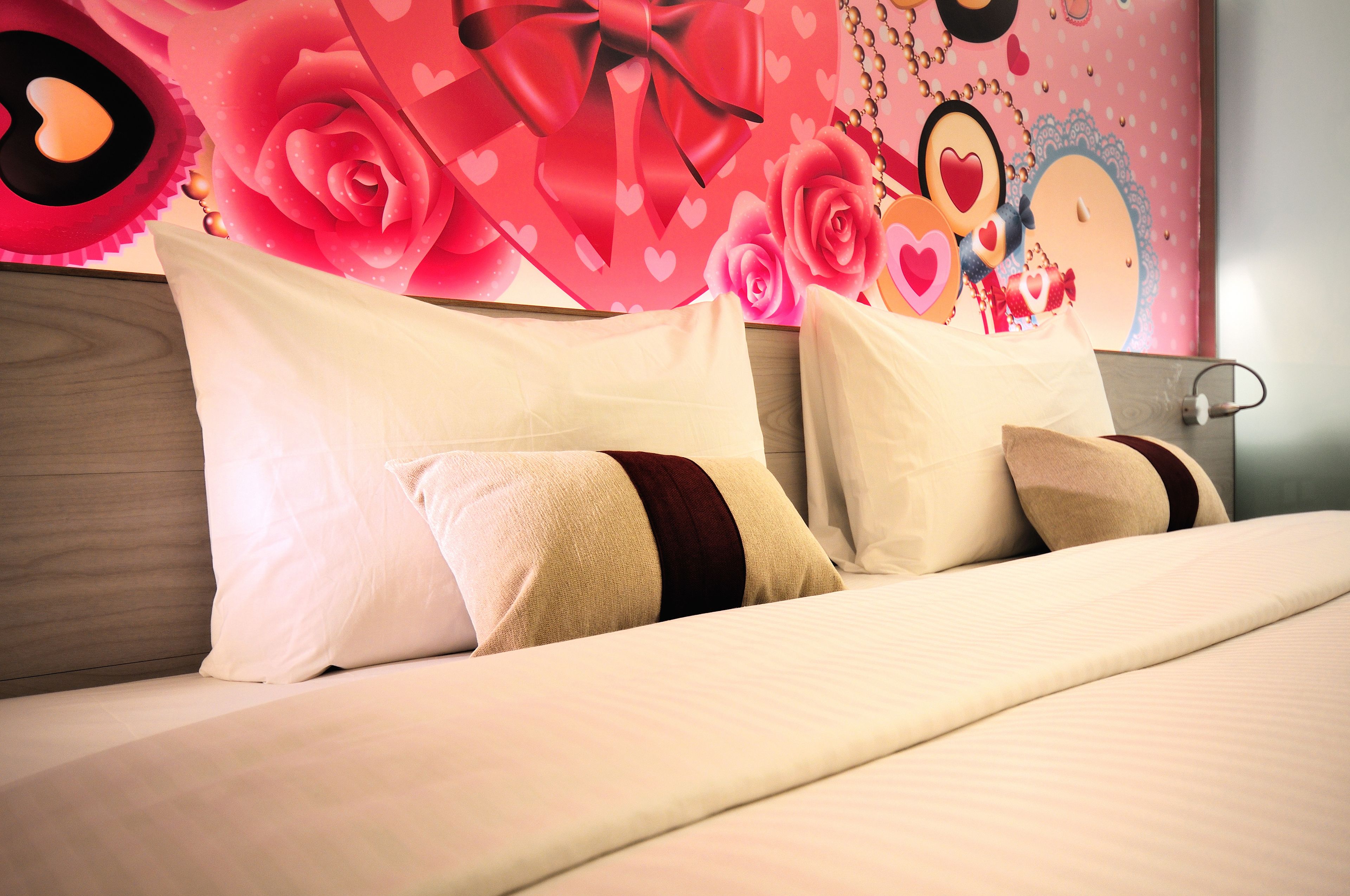 Een net opgemaakt bed met witte en beige kussens, een houten hoofdbord en een muur versierd met roze rozen en harten.